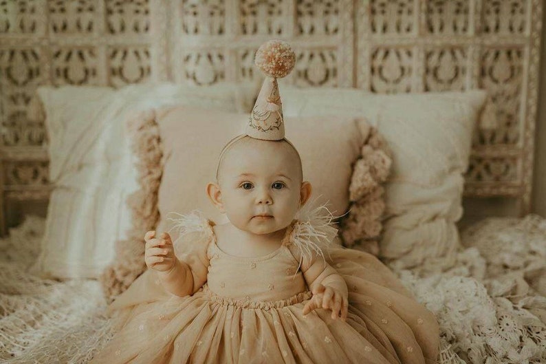 K&ouml;nnte beinhalten: Ein kleines M&auml;dchen in einem beigefarbenen T&uuml;llkleid mit wei&szlig;em Federbesatz und einer rosa-wei&szlig;en Partym&uuml;tze. Das Baby sitzt auf einem wei&szlig;en Bett mit einer wei&szlig;en H&auml;keldecke.