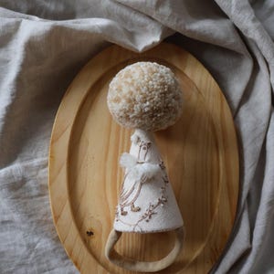 K&ouml;nnte beinhalten: Eine handgefertigte Puppe mit einem beigefarbenen Pomponkopf und einem wei&szlig;en Stoffkleid mit Stickereien. Die Puppe sitzt auf einem Holztablett.