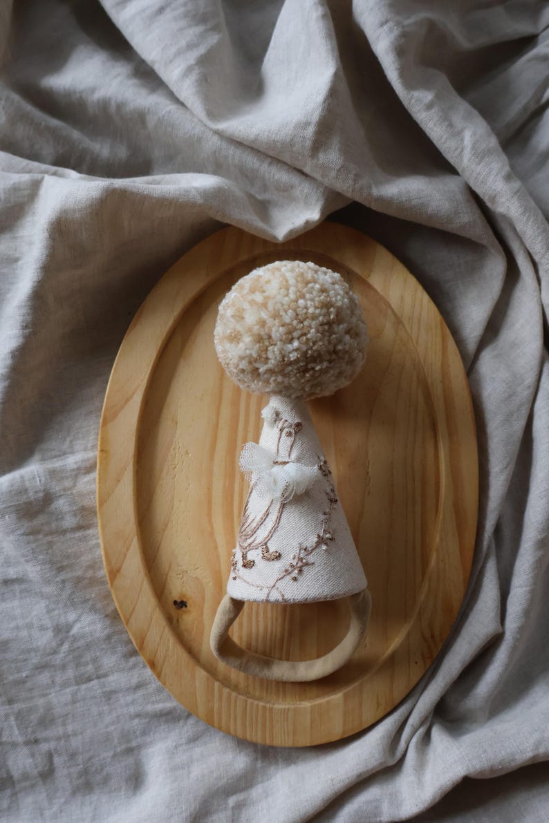 K&ouml;nnte beinhalten: Eine handgefertigte Puppe mit einem beigefarbenen Pomponkopf und einem wei&szlig;en Stoffkleid mit Stickereien. Die Puppe sitzt auf einem Holztablett.