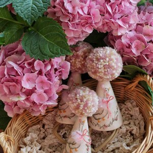K&ouml;nnte beinhalten: Drei handgefertigte Partyh&uuml;te in Form von G&auml;nsen mit rosa Bommeln und gestickten Details werden in einem Weidenkorb mit Spitzenfutter pr&auml;sentiert. Die H&uuml;te sind aus wei&szlig;em Stoff und haben rosa Schleifenakzente.