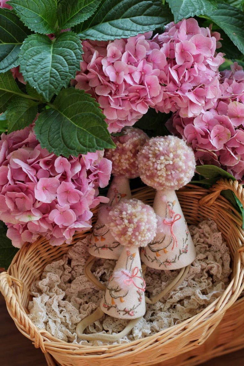 K&ouml;nnte beinhalten: Drei handgefertigte Partyh&uuml;te in Form von G&auml;nsen mit rosa Bommeln und gestickten Details werden in einem Weidenkorb mit Spitzenfutter pr&auml;sentiert. Die H&uuml;te sind aus wei&szlig;em Stoff und haben rosa Schleifenakzente.