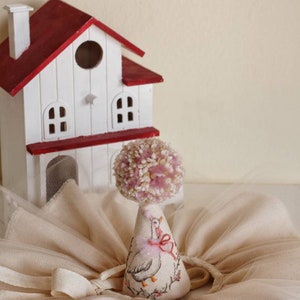K&ouml;nnte beinhalten: Ein wei&szlig;es und rotes Miniaturhaus mit einem rosa Bommel auf einem wei&szlig;en Kegel mit einem rosa Band und einem gestickten G&auml;nsemuster. Der Kegel steht auf einem wei&szlig;en T&uuml;llstoff.