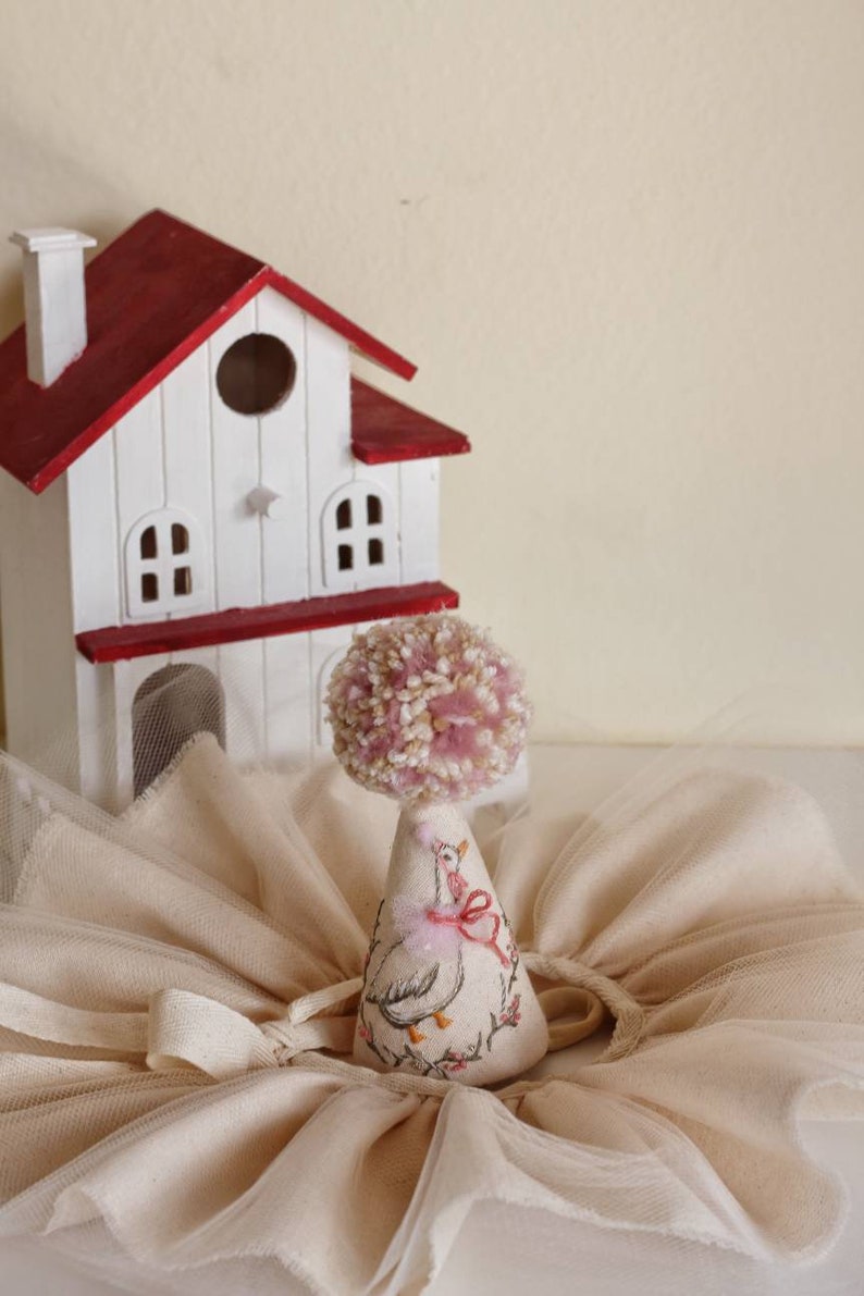K&ouml;nnte beinhalten: Ein wei&szlig;es und rotes Miniaturhaus mit einem rosa Bommel auf einem wei&szlig;en Kegel mit einem rosa Band und einem gestickten G&auml;nsemuster. Der Kegel steht auf einem wei&szlig;en T&uuml;llstoff.