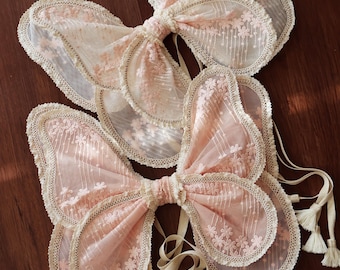 May include: Two pairs of delicate butterfly wings made of sheer fabric with lace trim. The wings are in shades of pink and cream and are tied together with satin ribbons.