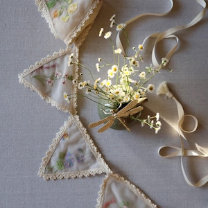 May include: A cream-colored fabric banner with embroidered floral designs and lace trim. The banner is composed of triangular pennants connected by a ribbon. A small vase with daisies and a gold dragonfly decoration is next to the banner.