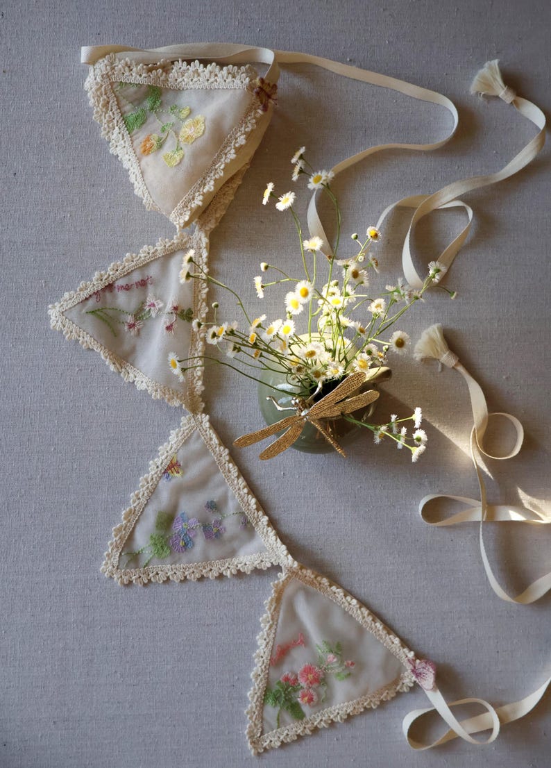 May include: A cream-colored fabric banner with embroidered floral designs and lace trim. The banner is composed of triangular pennants connected by a ribbon. A small vase with daisies and a gold dragonfly decoration is next to the banner.