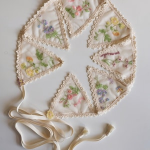 May include: A cream-colored floral banner with embroidered flowers in pink, yellow, and purple. Each triangular pennant is edged with lace and features a different flower design and text. A cream-colored ribbon is attached.