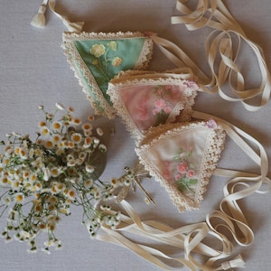 May include: Three triangular fabric sachets with floral embroidery and lace trim in shades of green, pink, and white. Each sachet has a cream-colored ribbon attached. A bouquet of white flowers and a decorative dragonfly are also present.