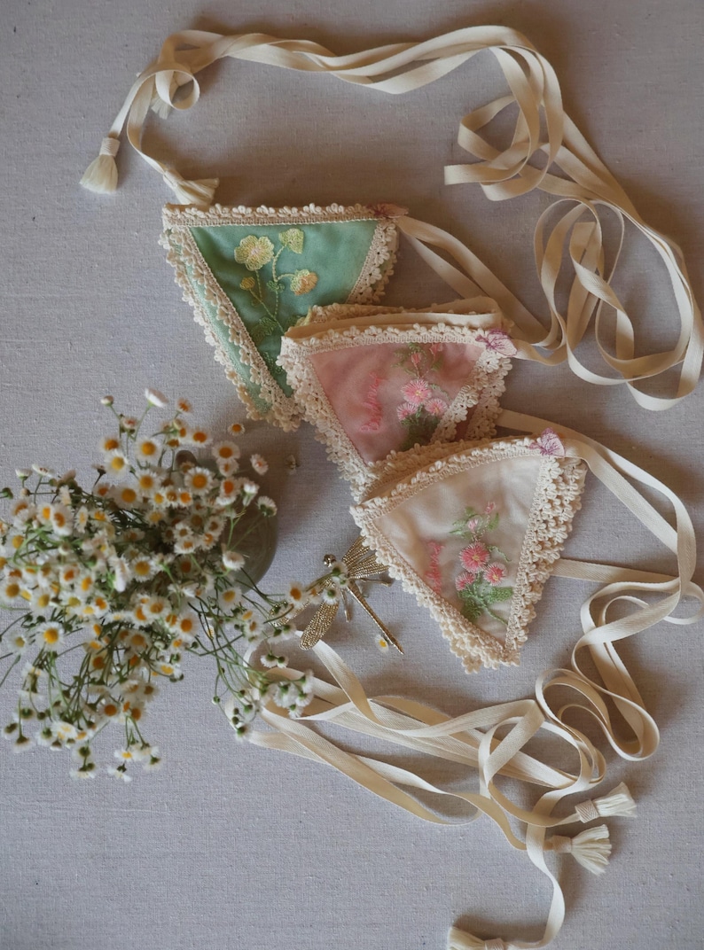 May include: Three triangular fabric sachets with floral embroidery and lace trim in shades of green, pink, and white. Each sachet has a cream-colored ribbon attached. A bouquet of white flowers and a decorative dragonfly are also present.
