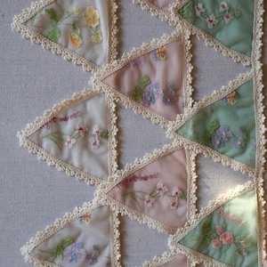 May include: Close-up of a textile with triangular patches in pastel colors. Each patch features embroidered flowers and the words "forget me not". The edges are trimmed with a delicate lace.