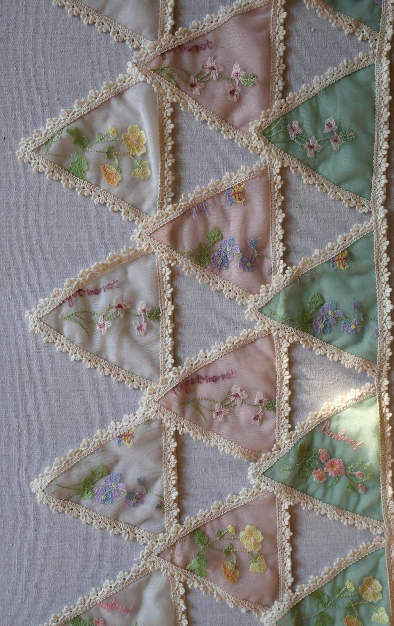 May include: Close-up of a textile with triangular patches in pastel colors. Each patch features embroidered flowers and the words "forget me not". The edges are trimmed with a delicate lace.