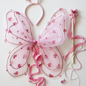 May include: Pink butterfly wings with a pink headband and a pink wand with a heart-shaped top. The wings are made of tulle and have a glitter heart pattern. The headband has two heart-shaped decorations. The wand has a wooden stick and a pink ribbon.