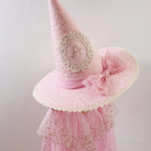 May include: A pink pointed hat featuring a gold star pattern, a cream-colored lace trim, and a large matching bow. The front of the cone displays a circular embroidered patch with a decorative lace border and the letter A stitched in the center.