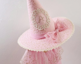 May include: A pink pointed hat featuring a gold star pattern, a cream-coloured lace trim, and a large matching bow. The front of the cone displays a circular embroidered patch with a decorative lace border and the letter A stitched in the centre.