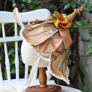 May include: A tan felt bonnet featuring a pointed top with a knotted tip and a wide, flared brim. The fabric is decorated with an embroidered pattern of brown and grey branches. A cluster of artificial autumn flowers sits on the side.