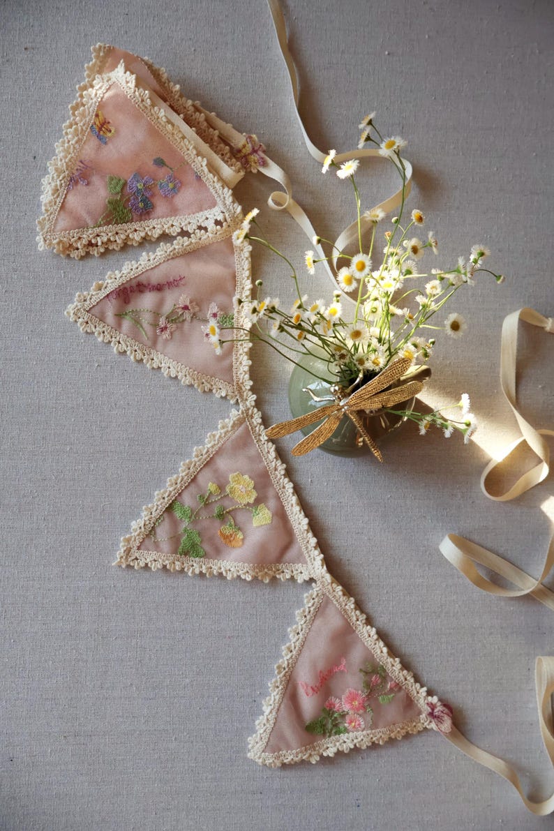 May include: A pink fabric banner with embroidered floral designs and lace trim. The banner is composed of triangular pennants, each featuring different flower patterns. A small vase with daisies and a gold dragonfly ornament is next to the banner.