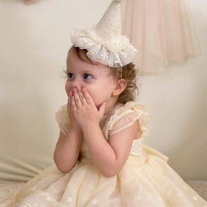 Peut inclure: Un jeune enfant portant un chapeau de fête en dentelle blanche avec un pompon blanc sur le dessus. L'enfant porte une robe jaune avec des bordures en dentelle blanche et se couvre la bouche avec les mains.