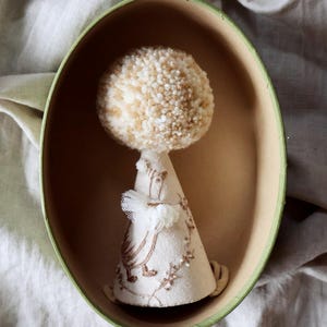 K&ouml;nnte beinhalten: Ein beige-wei&szlig;er Pompon sitzt auf einer kegelf&ouml;rmigen Stoffpuppe mit Stickereien. Die Puppe befindet sich in einer ovalen gr&uuml;n-braunen Box.