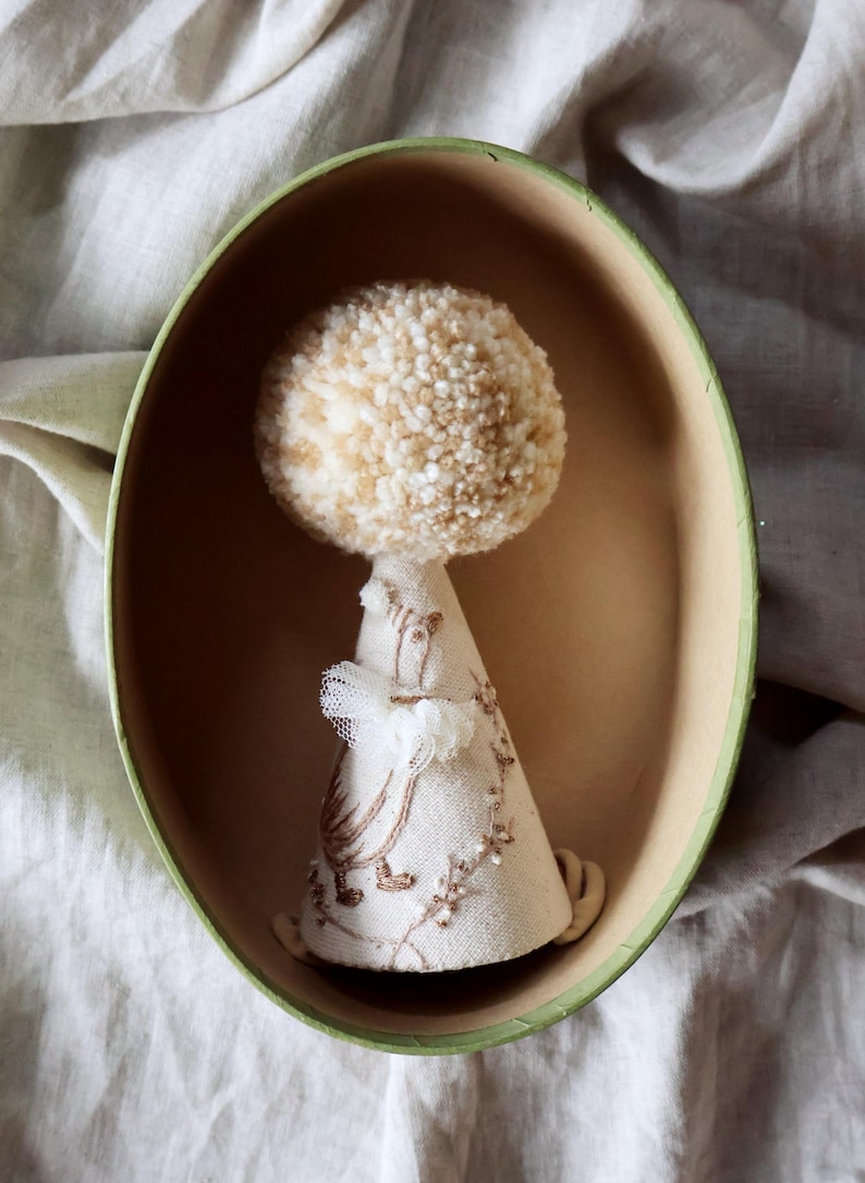 K&ouml;nnte beinhalten: Ein beige-wei&szlig;er Pompon sitzt auf einer kegelf&ouml;rmigen Stoffpuppe mit Stickereien. Die Puppe befindet sich in einer ovalen gr&uuml;n-braunen Box.