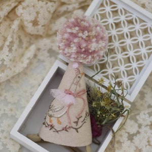 K&ouml;nnte beinhalten: Ein rosa-wei&szlig;er Pompon sitzt auf einem handgefertigten Stoffkegel mit gestickten Details eines Vogels und einer Schleife. Der Kegel befindet sich in einer wei&szlig;en Box mit einem dekorativen Gittermuster.