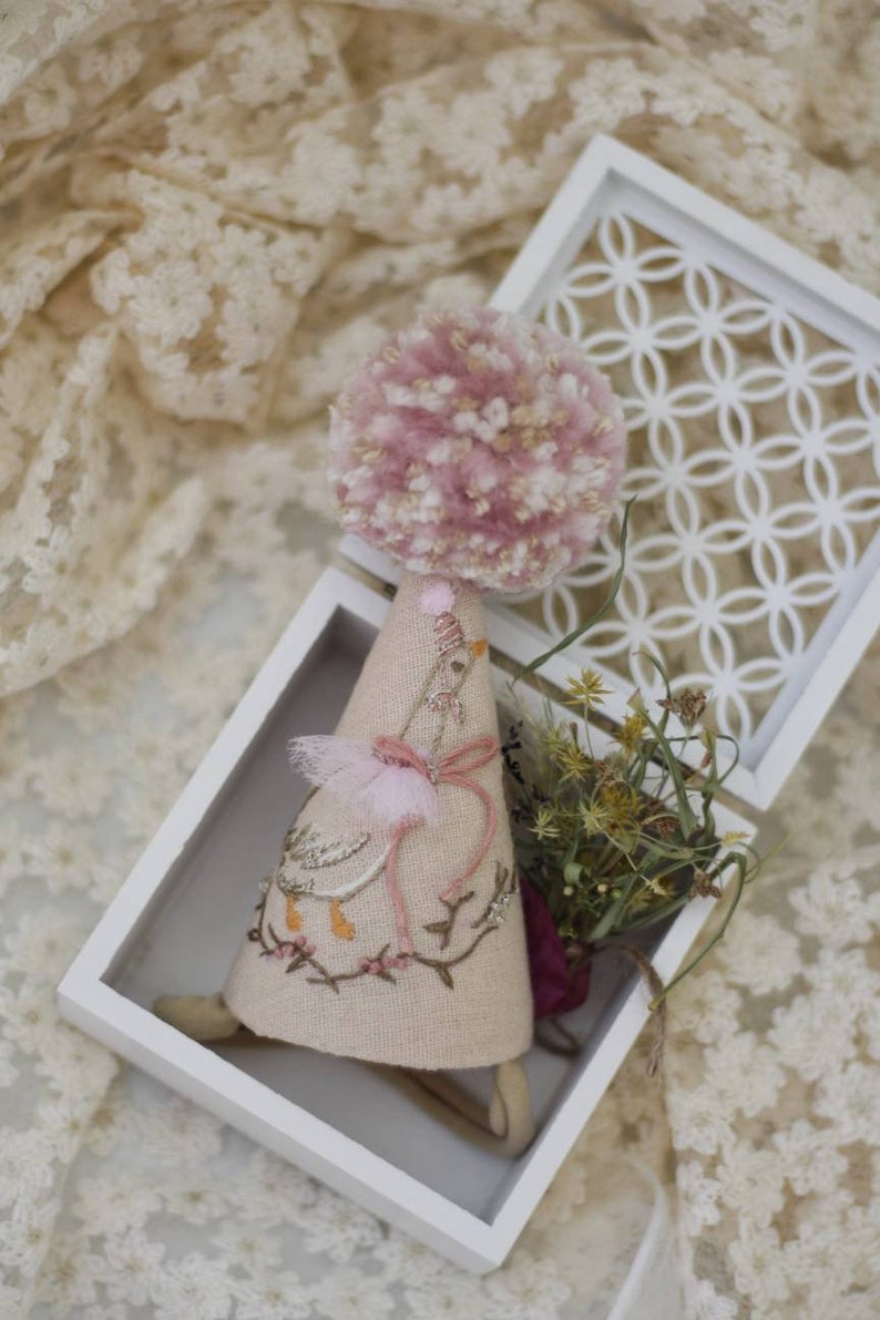 K&ouml;nnte beinhalten: Ein rosa-wei&szlig;er Pompon sitzt auf einem handgefertigten Stoffkegel mit gestickten Details eines Vogels und einer Schleife. Der Kegel befindet sich in einer wei&szlig;en Box mit einem dekorativen Gittermuster.