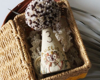 May include: A cream-coloured party hat with a lion design and the name "Austin" embroidered on it sits in a woven basket. The hat has a brown pom-pom and the number "3" embroidered on it. The basket is light brown.