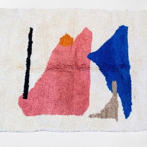 May include: A cream-colored, rectangular rug with an abstract design. The rug features a large pink shape, a blue triangle, a black vertical line, and an orange circle. The rug has white fringe on the edges.