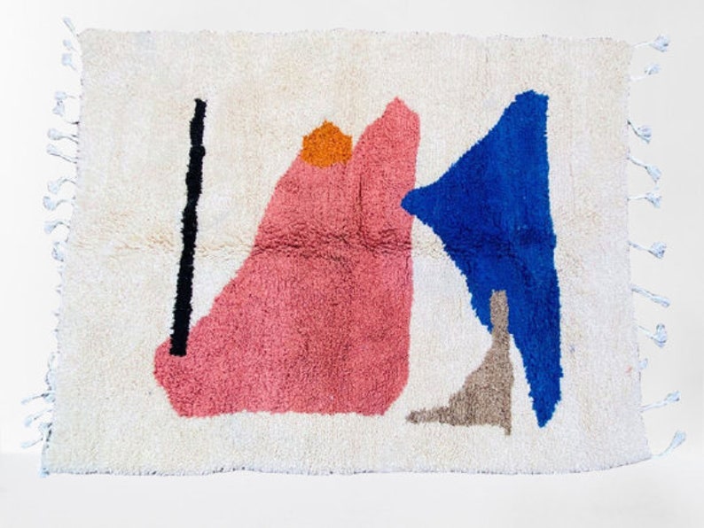 May include: A cream-colored, rectangular rug with an abstract design. The rug features a large pink shape, a blue triangle, a black vertical line, and an orange circle. The rug has white fringe on the edges.