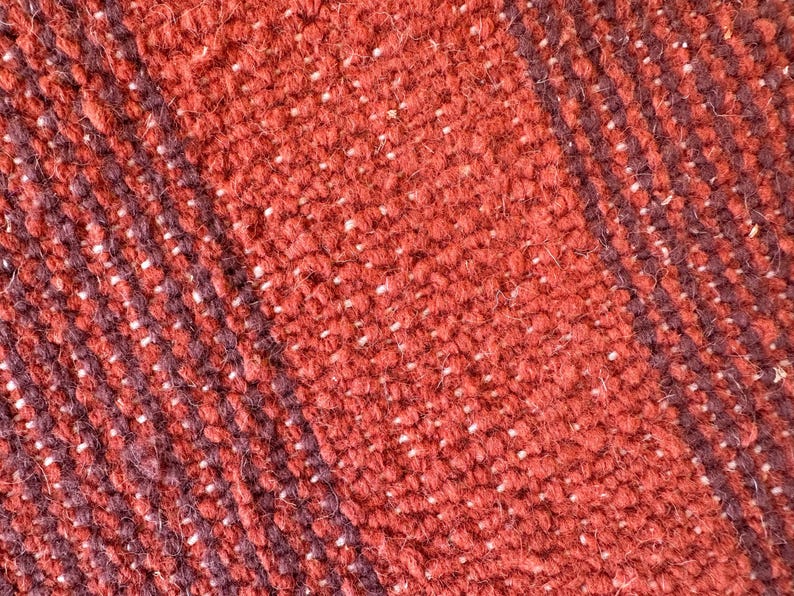 May include: Close-up of a woven fabric with a red and dark brown striped pattern. The fabric has a textured, fuzzy surface.
