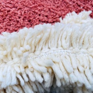 May include: Close-up of a textured rug with a cream-colored fringe and a dusty rose background. The fringe is thick and shaggy, with visible stitching. The rug's texture is soft and inviting, suggesting a cozy home decor item.