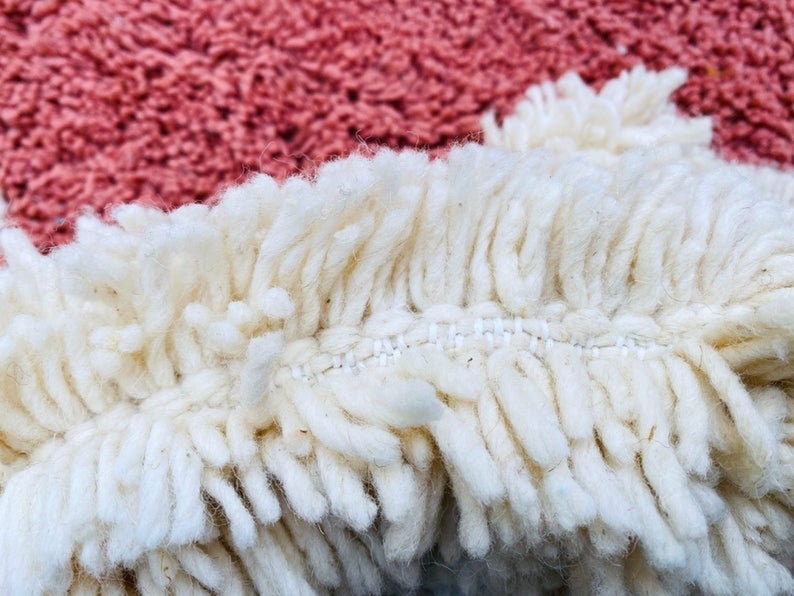 May include: Close-up of a textured rug with a cream-colored fringe and a dusty rose background. The fringe is thick and shaggy, with visible stitching. The rug's texture is soft and inviting, suggesting a cozy home decor item.
