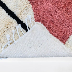 May include: A close-up of a textured rug with a geometric design. The rug features a cream-colored background with a large pink heart shape and a black diagonal stripe. The rug has white fringe.