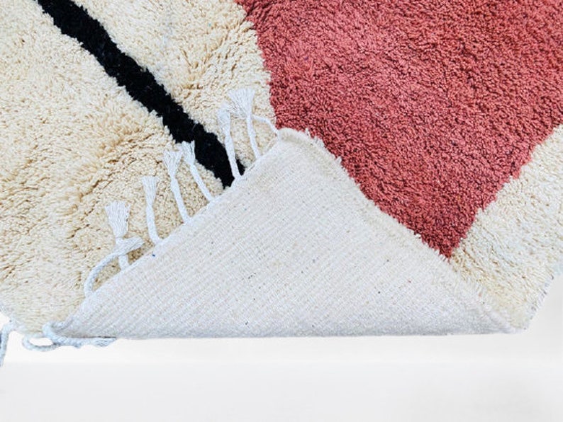 May include: A close-up of a textured rug with a geometric design. The rug features a cream-colored background with a large pink heart shape and a black diagonal stripe. The rug has white fringe.