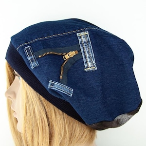 Op de afbeelding: Een donkerblauwe denim beanie-muts met een rits en nepzakken. De muts heeft een donkerblauwe geribbelde band en een bruine suède-achtige onderkant. De rits is goudkleurig.
