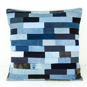 May include: Square decorative pillow with a patchwork design made from various shades of blue denim. The pillow features a mix of light, medium, and dark blue denim pieces arranged in horizontal rows. The pillow is a square shape.
