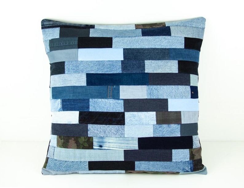 May include: Square decorative pillow with a patchwork design made from various shades of blue denim. The pillow features a mix of light, medium, and dark blue denim pieces arranged in horizontal rows. The pillow is a square shape.