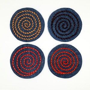 Peut inclure: Quatre dessous de verre circulaires en jean disposés en carré. Chaque dessous de verre présente un motif de broderie en spirale distinct avec des fils de couleurs contrastées : un jaune, un marron foncé et deux rouge vif avec des accents blancs.