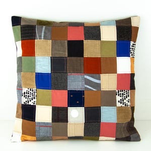 Peut inclure: Coussin carré en patchwork avec une variété de carrés de tissu dans les tons de marron, bleu, rouge et vert. Le coussin a un fond neutre et une fermeture à bouton. Un élément de décoration pour la maison.