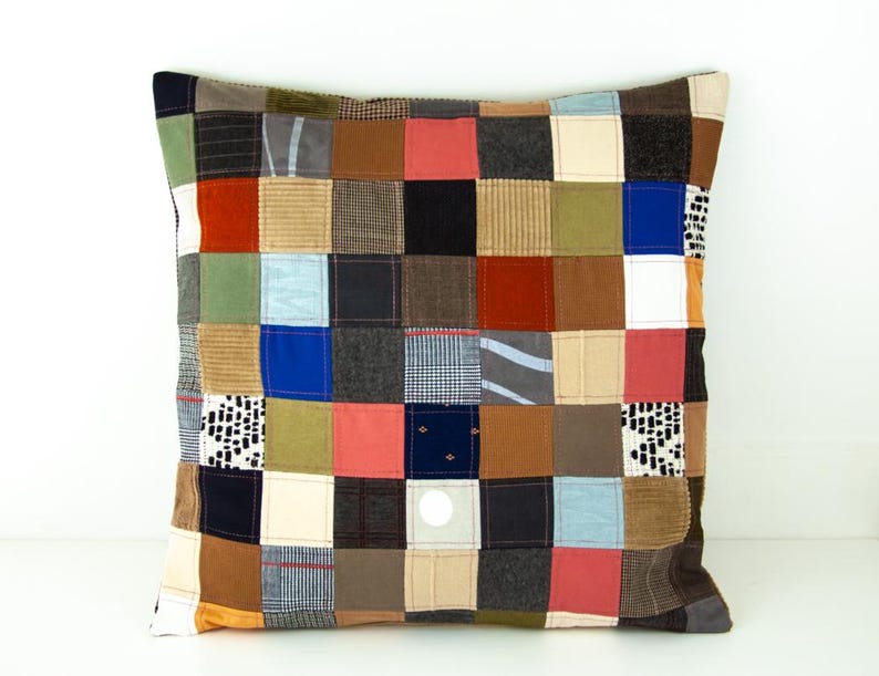 Puede incluir: Coj&iacute;n cuadrado de patchwork con una variedad de cuadrados de tela en tonos marrones, azules, rojos y verdes. El coj&iacute;n tiene un fondo neutro y un cierre de bot&oacute;n. Un elemento decorativo para el hogar.