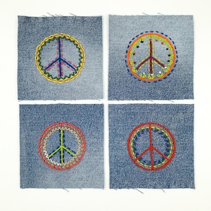 Könnte beinhalten: Vier Jeansstoff-Patches, jeweils mit einem farbenfrohen, gestickten Friedenszeichen. Die Zeichen sind in verschiedenen Farben gehalten, darunter Gelb, Orange, Rot und Blau, mit detaillierten Stickereien. Die Patches sind quadratisch und haben einen rohen Rand.