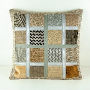Può includere: Un cuscino quadrato con un design a patchwork. Il cuscino è realizzato con diversi tessuti in tonalità di marrone, beige e grigio. I tessuti sono disposti a griglia, con ogni quadrato che presenta una texture o un motivo diverso.