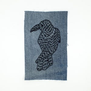 May include: A denim patch featuring a black embroidered raven design. The raven is depicted in profile, with intricate stitching detailing its feathers and form. The patch has a rectangular shape with frayed edges, giving it a vintage look.