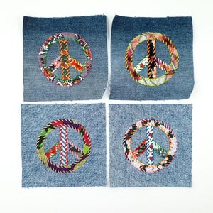 Peut inclure: Quatre écussons en denim bleu, chacun orné d'un symbole de paix coloré. Les symboles de paix sont faits de tissus à motifs variés, rouges, oranges, verts et violets, cousus sur des carrés de denim.