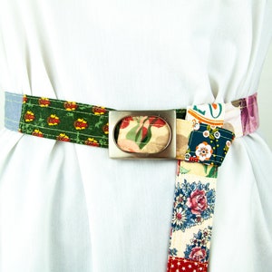 Peut inclure: Une ceinture en tissu patchwork colorée avec une boucle ovale argentée. La ceinture est nouée à l'avant et présente une variété de motifs floraux et géométriques.
