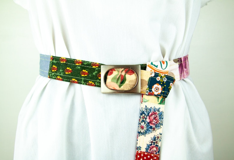 Op de afbeelding: Een kleurrijke patchwork stoffen riem met een zilveren ovale gesp. De riem is aan de voorkant in een knoop geknoopt en heeft verschillende bloem- en geometrische patronen.