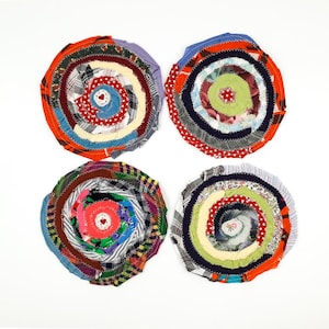 May include: Four handmade, circular coasters with a spiral design. Each coaster is made from fabric scraps in various colors, including red, blue, green, and white. The coasters are arranged on a white background.