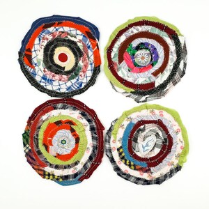 May include: Four handmade, circular coasters with a spiral design. Each coaster is made from fabric scraps in various colours and patterns, including red, blue, green, and white. The coasters are arranged on a white background.