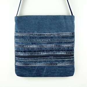 Peut inclure: Un sac bandoulière de forme carrée fabriqué en tissu denim bleu délavé moyen. Le panneau avant présente un motif patchwork horizontal composé de multiples bandes de denim superposées avec des coutures visibles et une fine bandoulière bleu foncé.