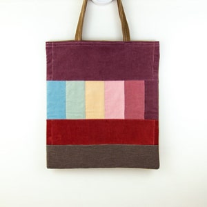 Puede incluir: Una bolsa de mano de pana color burdeos con un diseño de patchwork de pana azul pastel, verde, amarillo, rosa y rojo. La bolsa tiene asas de pana marrón y una base de pana marrón.