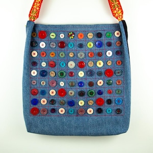 Peut inclure: Un sac fourre-tout en jean orné d'une grille de boutons colorés de différentes tailles et couleurs. Les boutons sont cousus sur le sac avec du fil rouge. Le sac a une sangle décorative avec un motif floral.